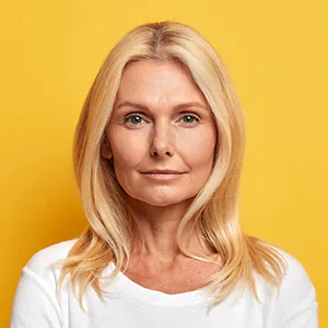 Woman with blonde hair wearing a white shirt, standing against a yellow background, with a neutral expression