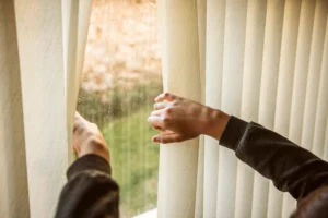 Person adjusting vertical blinds to reveal a view outside, controlling natural light and enhancing privacy in a cozy interior.
