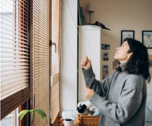 Person adjusting window blinds in a cozy home setting, showcasing Blinds as a provider of high-quality window treatments for privacy, light control, and style.