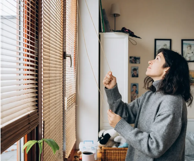 Person adjusting window blinds in a cozy home setting, showcasing Blinds as a provider of high-quality window treatments for privacy, light control, and style.